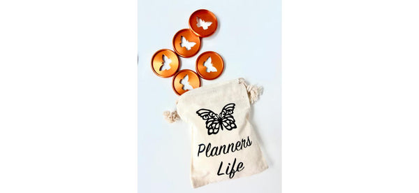 Five bronze butterfly-cutout planner binding discs above a cream drawstring pouch printed with a black butterfly and the words Planners Life