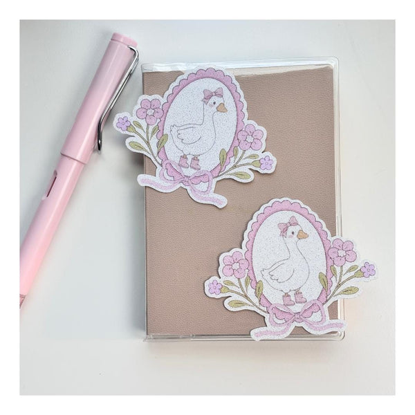 Two coquette goose die-cut stickers with pink floral wreaths and bows on a notebook, next to a pink pen