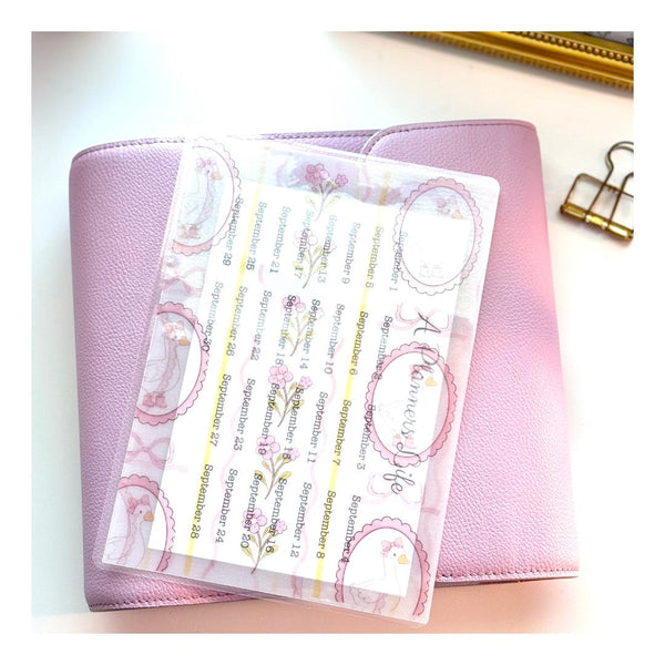 Close-up of a coquette goose vellum sticker sheet with September date labels, propped against a pink pouch beside a gold binder clip