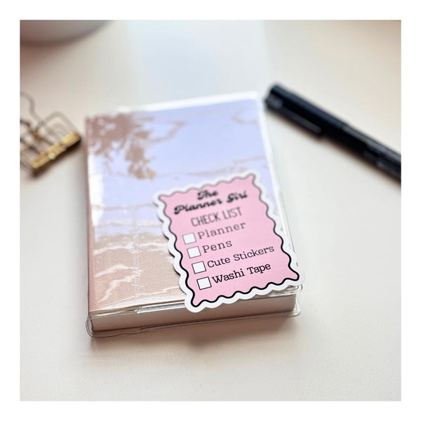 Planner girl checklist sticker on a marble-look planner cover with paperclip and pen
