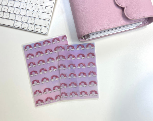 Two rainbow and cloud vellum sticker sheets on a white desk beside a closed pink scalloped-flap planner and a keyboard