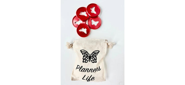 Five glossy red planner binding discs with white butterfly cutouts above a cream drawstring pouch printed with a black butterfly and the words Planners Life