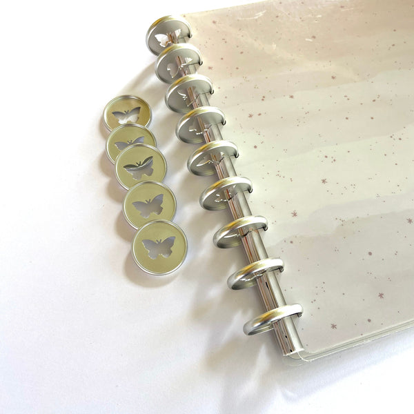 Close-up comparing five loose silver butterfly-cutout planner discs beside a metal binding rod threaded through a notebook to show how the disc mechanism works