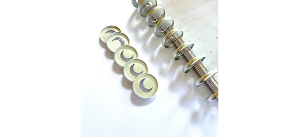 Five silver 32mm planner binding discs with crescent moon cutouts lined up beside the disc-bound spine of a cream speckled notebook