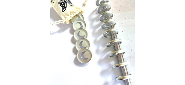 Close-up of five loose silver crescent moon planner binding discs next to a notebook's disc-bound spine, with a cream butterfly-print pouch above