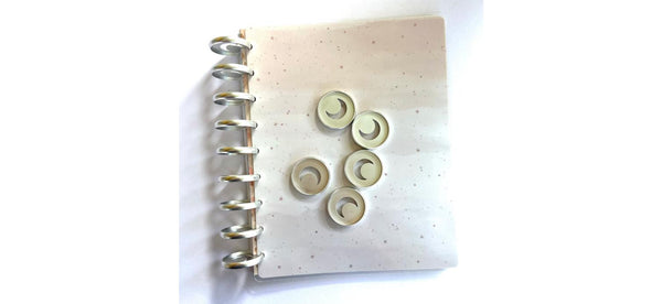 Five silver crescent moon cutout planner discs arranged on the cream speckled cover of a silver disc-bound notebook