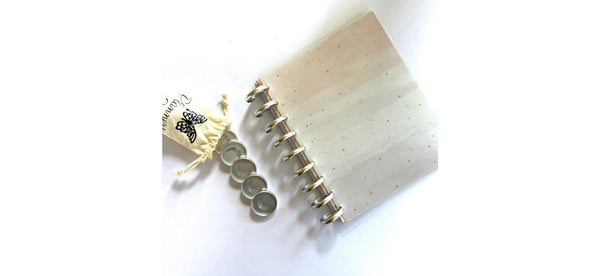 Silver crescent moon planner binding discs stacked beside a cream drawstring storage pouch with a butterfly logo, resting on a disc-bound cream speckled notebook
