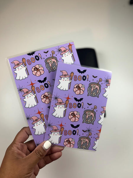 A hand holding two purple pocket folders printed with ghosts in witch hats, BOO text, bats, and jack-o'-lanterns
